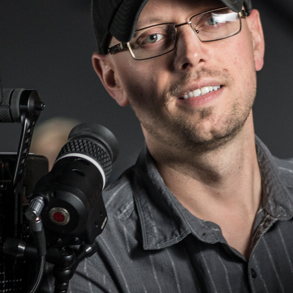 A close-up shot of Adrian's face, wearing a black baseball cap and black button-up shirt, holding a camera off to the side of the frame. 