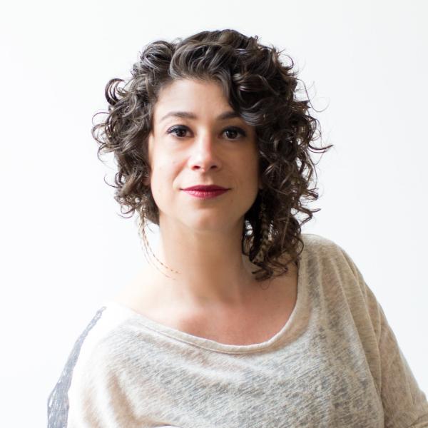 headshot of a white woman with curly brown hair and red lipstick, wearing a cream-colored sweater and looking directly at the camera.