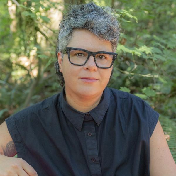 A white genderqueer person with short salt and pepper hair and black glasses looks gently into the camera.