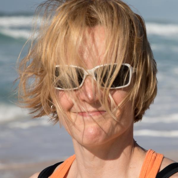 Photo of Claire Jones, a white person with chin-length blond hair at the beach. Clair's hair is blown by the breeze, pushing the front of it onto their face.