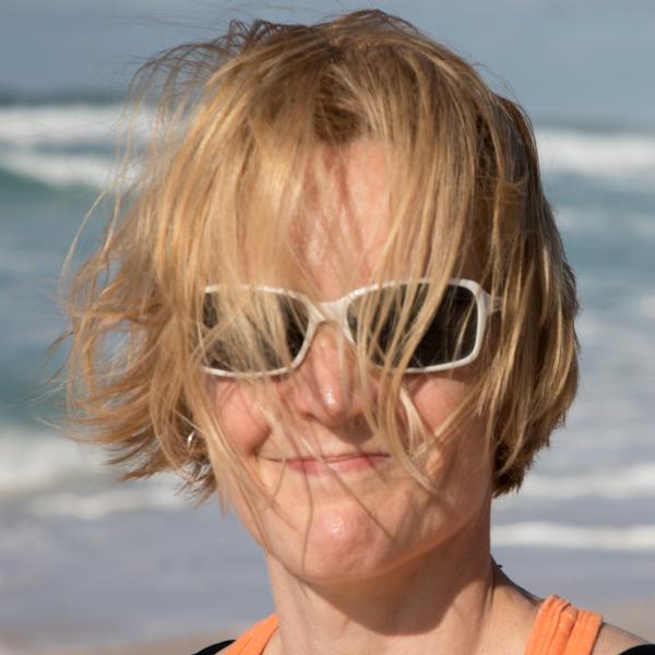 Photo of Claire Jones, a white person with chin-length blond hair at the beach. Clair's hair is blown by the breeze, pushing the front of it onto their face.