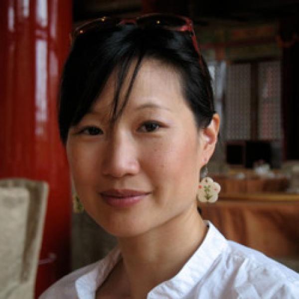 A headshot of filmmaker Debbie Lum smiling softly towards the camera. Her hair is tied back and her side bangs fall across her forehead. She is wearing a white collared shirt, red sunglasses, and flower-shaped earrings.