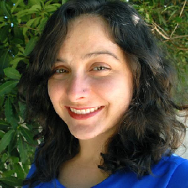 A headshot of the filmmaker Kimberly Bautista outside near a lush green tree. She has dark wavy hair, a small nose piercing and a broad smile, and she wears a blue shirt.
