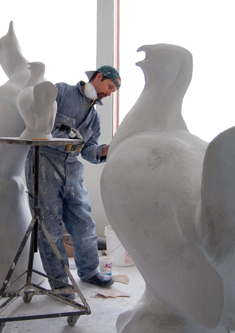 An artist in blue coveralls and a baseball cap surrounded by sculptures of birds. He is focused on one of them. They are all white and some are bigger than him. The room is all white and large windows show nothing but white light.