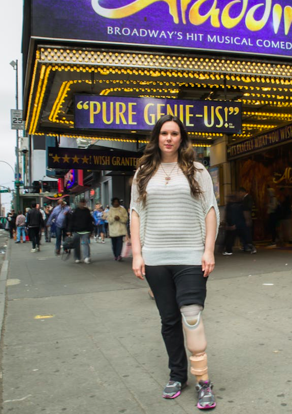 A white woman with long brown hair, a striped white and beige top, black pants and running shoes, standing on a city sidewalk in front of the awning for the theatrical production of Aladdin. She has her left pant leg hiked up to display her above-knee prosthetic leg.