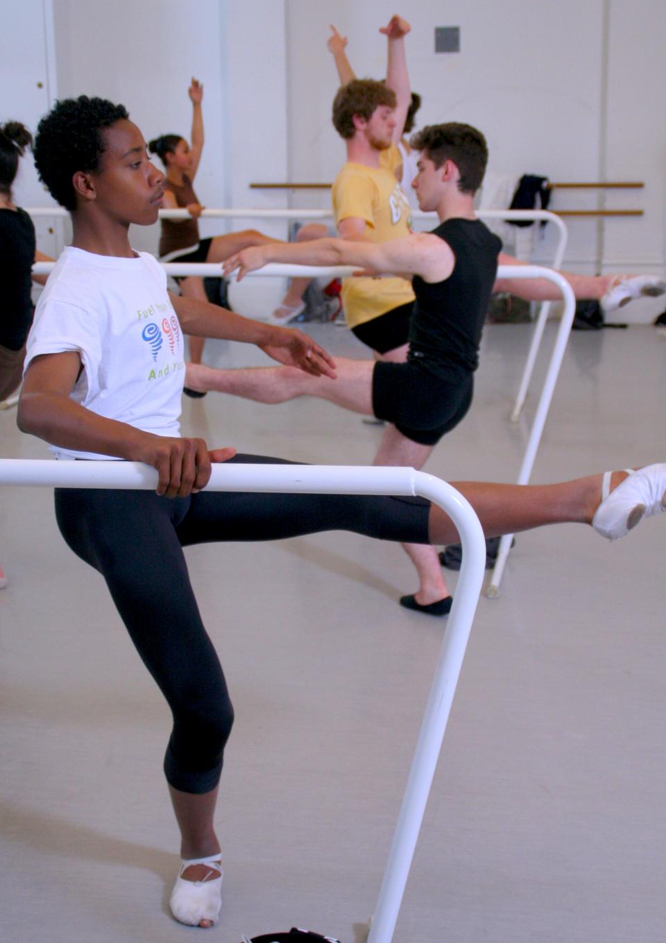Young ballet dancers of different genders and ethnicities are performing stretches in a dance room. They are holding white bars, and their legs and arms are extended. They wear white ballet shoes.
