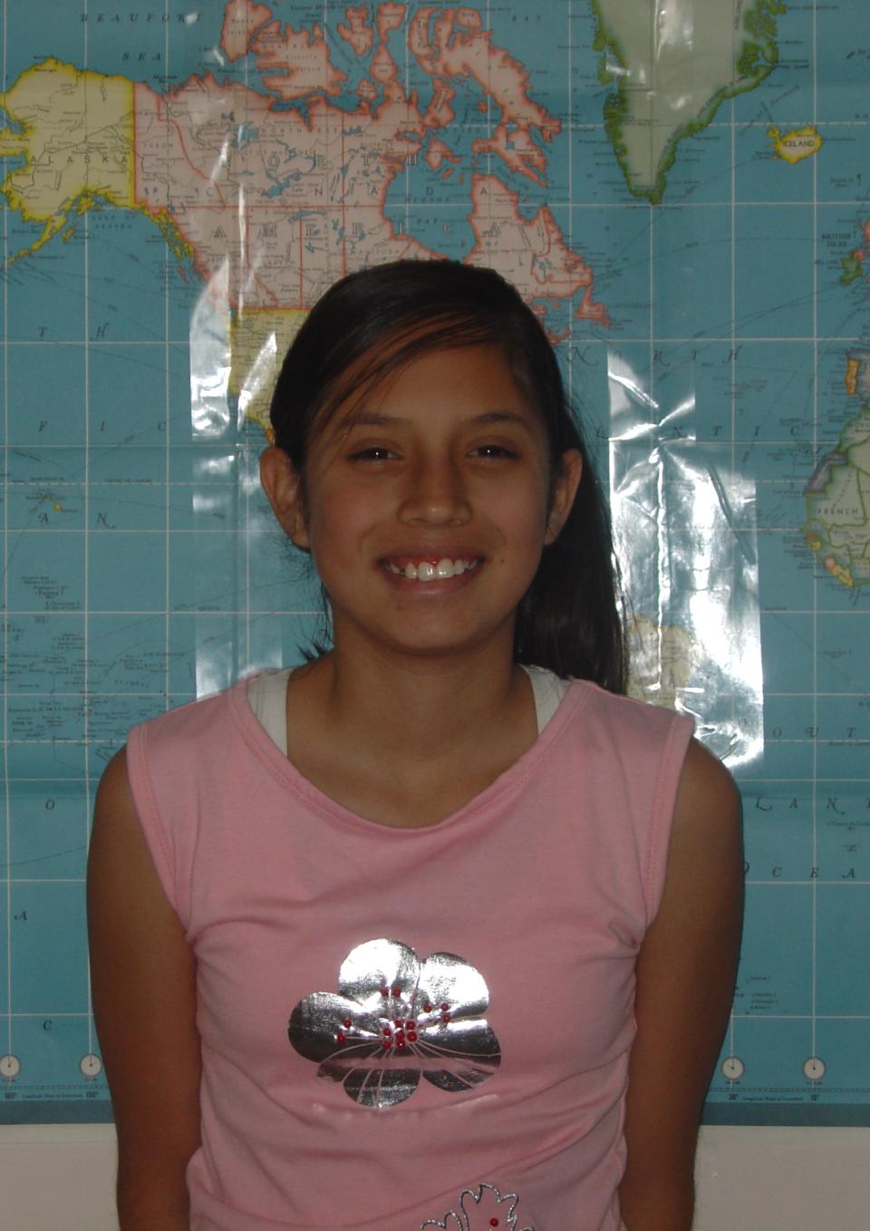 An adolescent Latina girl wearing a pink t-shirt with a silver flower on it stands in front of a large map of the world, smiling, her arms at her sides.