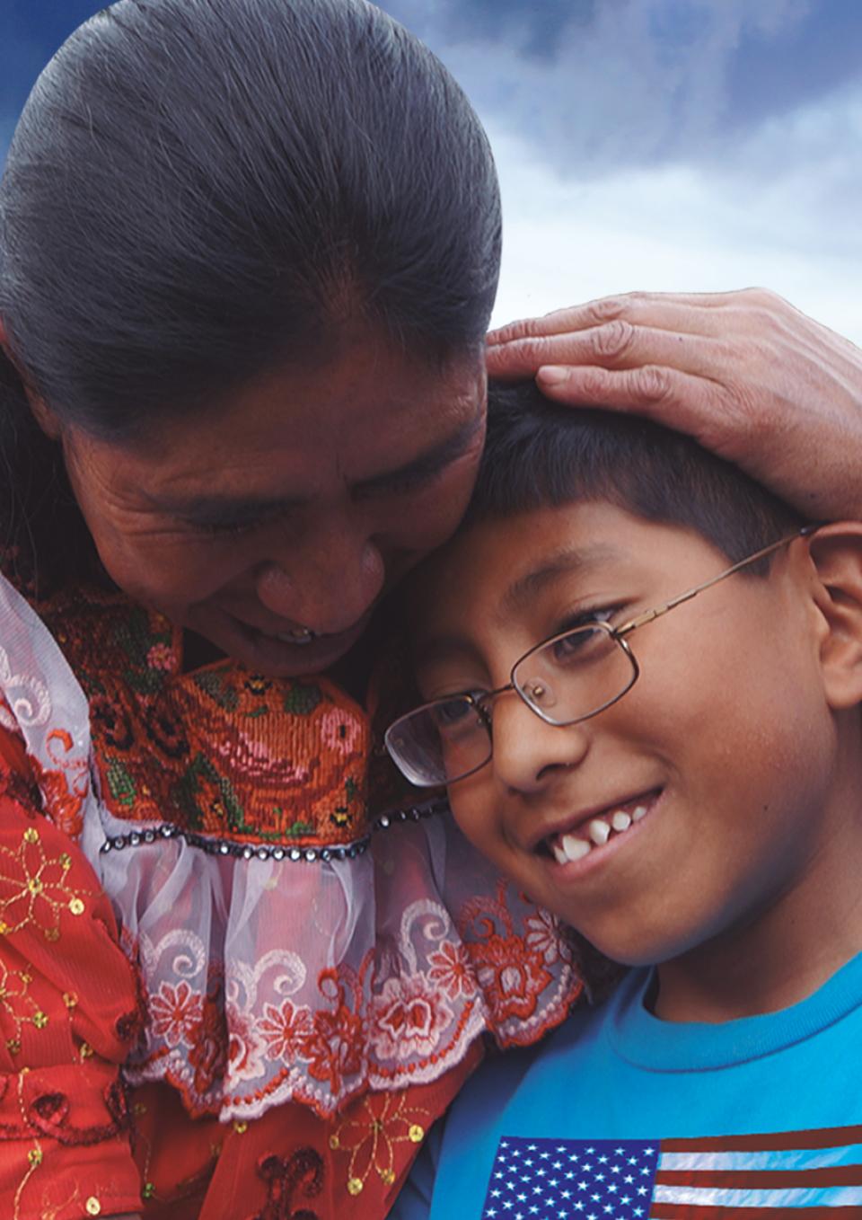 In Guatemala, a woman cradles the head of a young person. They are both smiling. The woman wears a red traditional Guatemalan dress and the boy wears a blue t-shirt with a U.S. flag printed on it.