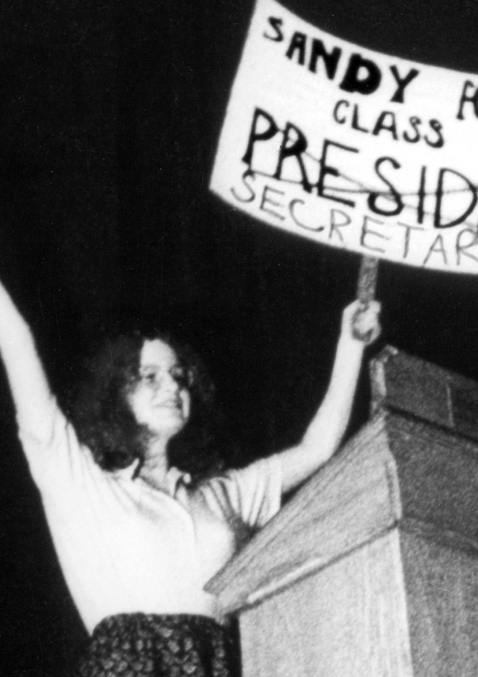 In this black and white still from the film, a young woman triumphantly holds up a sign that reads “Sandy For Class President.” The word ‘President’ is crossed out and underneath is written “Secretary”. She’s onstage at a lectern.