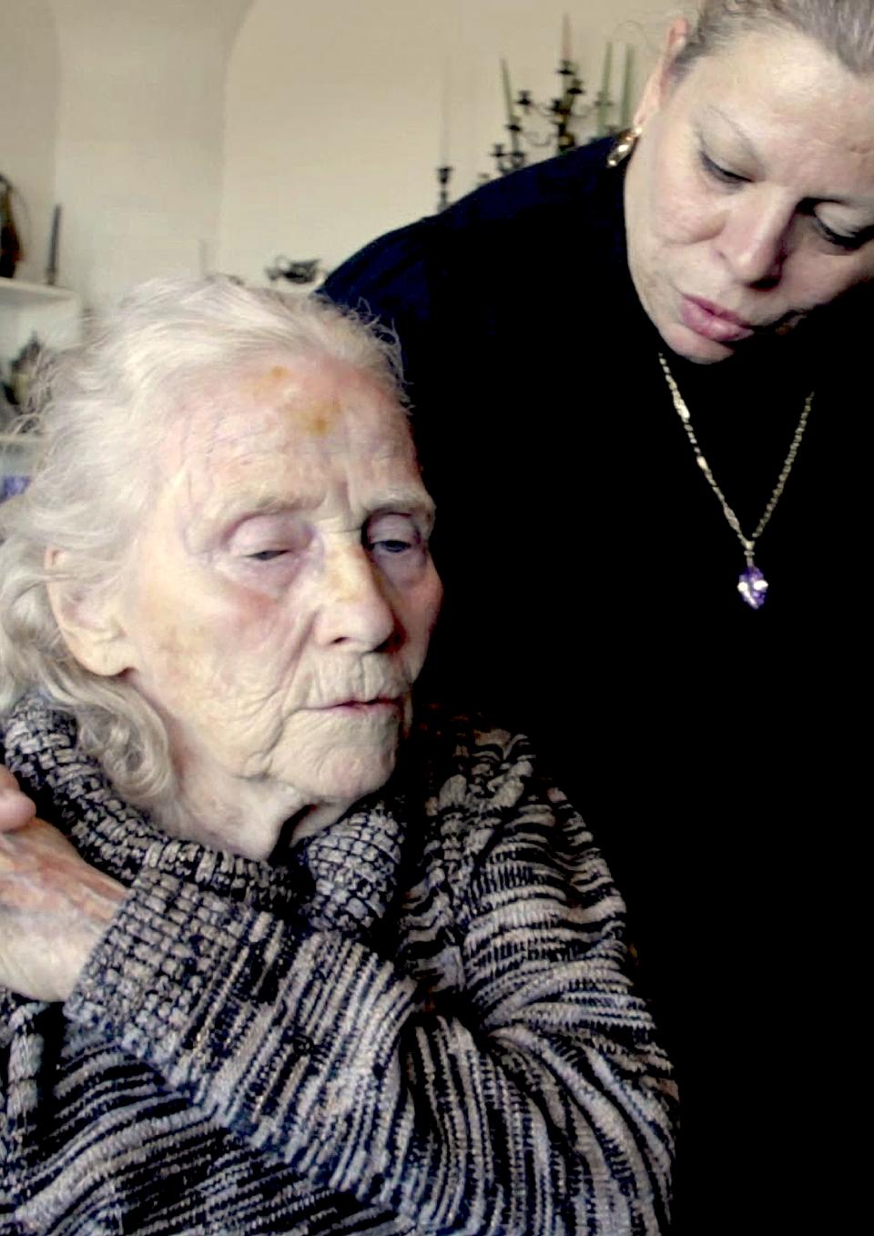 A middle aged woman is standing over an elderly woman with her arm around her. 