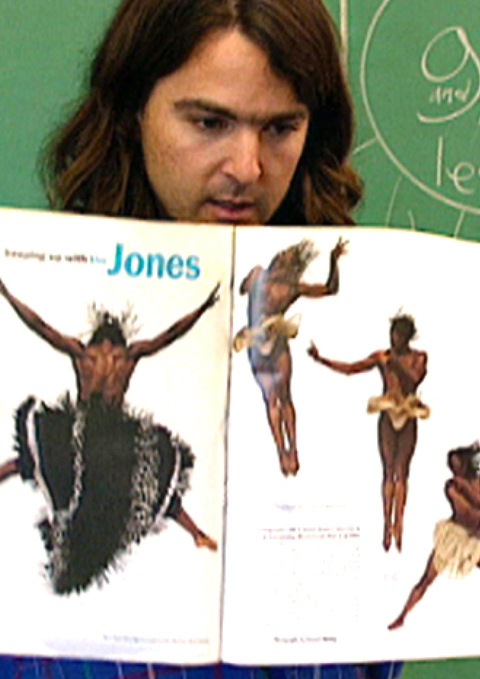 A teacher with brown hair stands in front of a chalkboard holding open a book with pictures of acrobatic Black dancers in fabulous costumes. Behind her is a chalkboard with the words gay and/or lesbian inside a circle with lines pointing out of it like a sun.