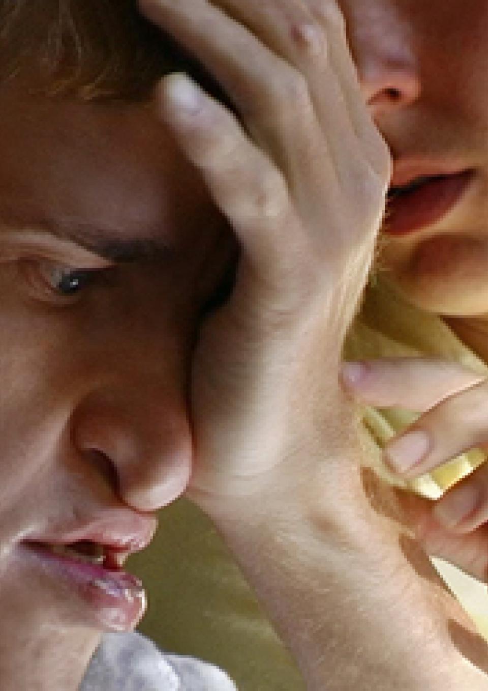 A young white man with multiple disabilities rests his palm against his forehead as a white woman reaches to touch his hand.