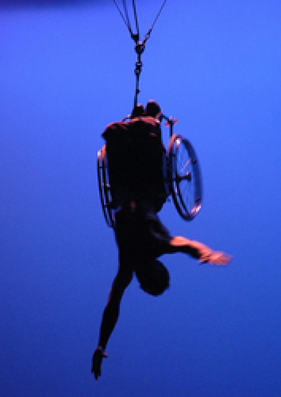 A person in a wheelchair suspended upside down with their arms extended, against a bright blue background.