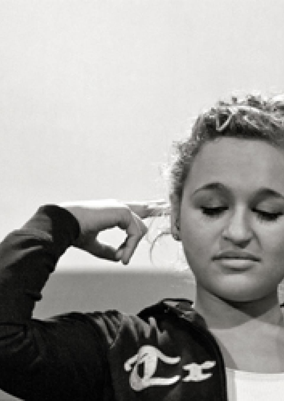 A black and white still from the New Day film Deaf Jam. Young ASL poet Aneta Brodski closes her eyes with a vulnerable but dignified expression on her face. She holds out one finger pointed out towards her ear. She wears her curly hair up in a bun and her hoodie is open to show a white top underneath.