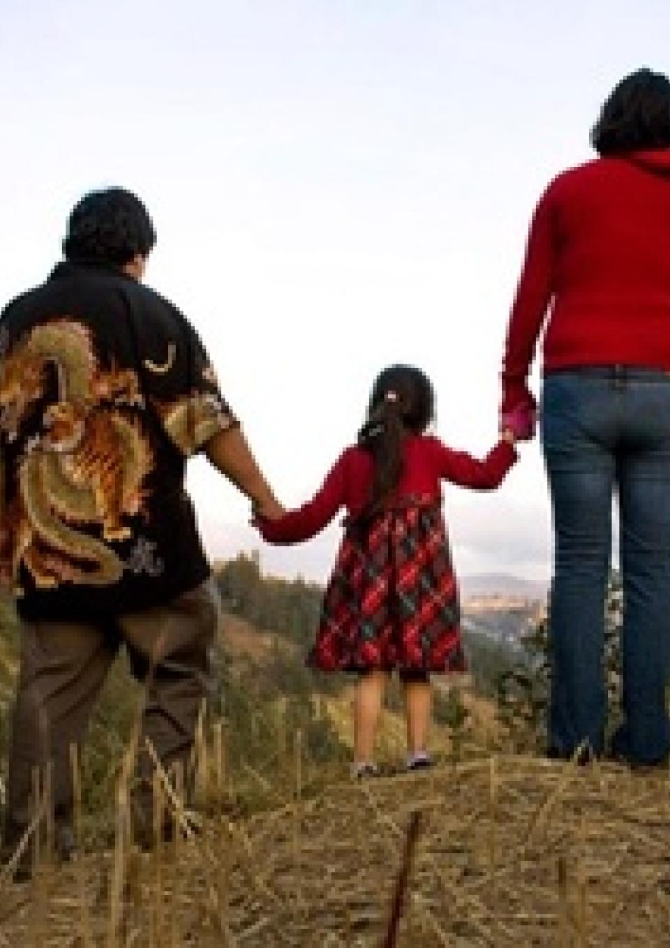 A still from the New Day film Sin Pais. The backs of three family members holding hands, a small child in the middle. They walk on a sandy path with tall grass on either side.