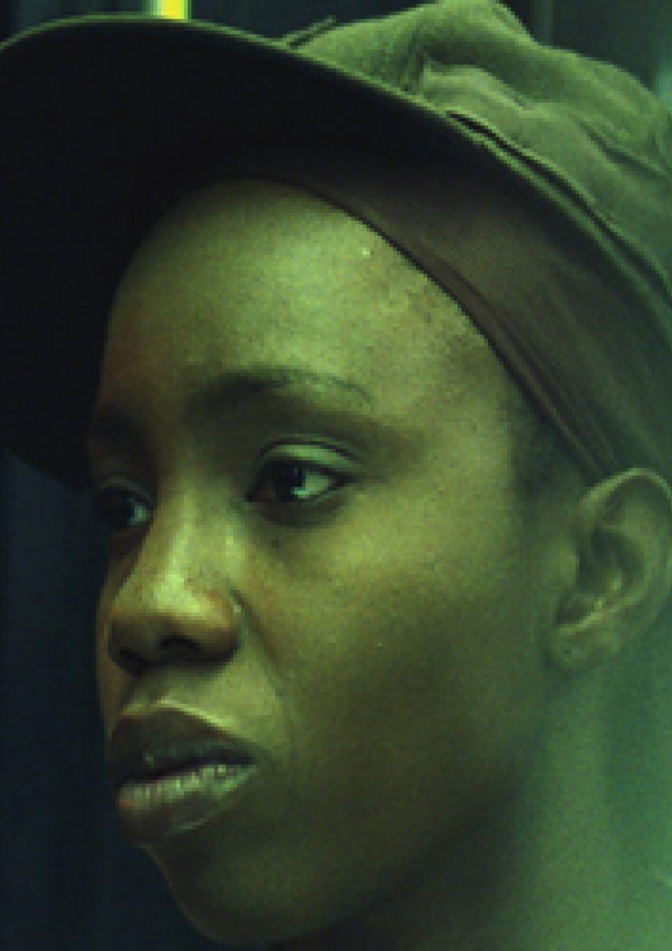 A close up image of a Black youth sitting in a bus with their face reflected on the bus window as they peer out into the distance. A harsh white street light obscures their reflection against the dark night background.