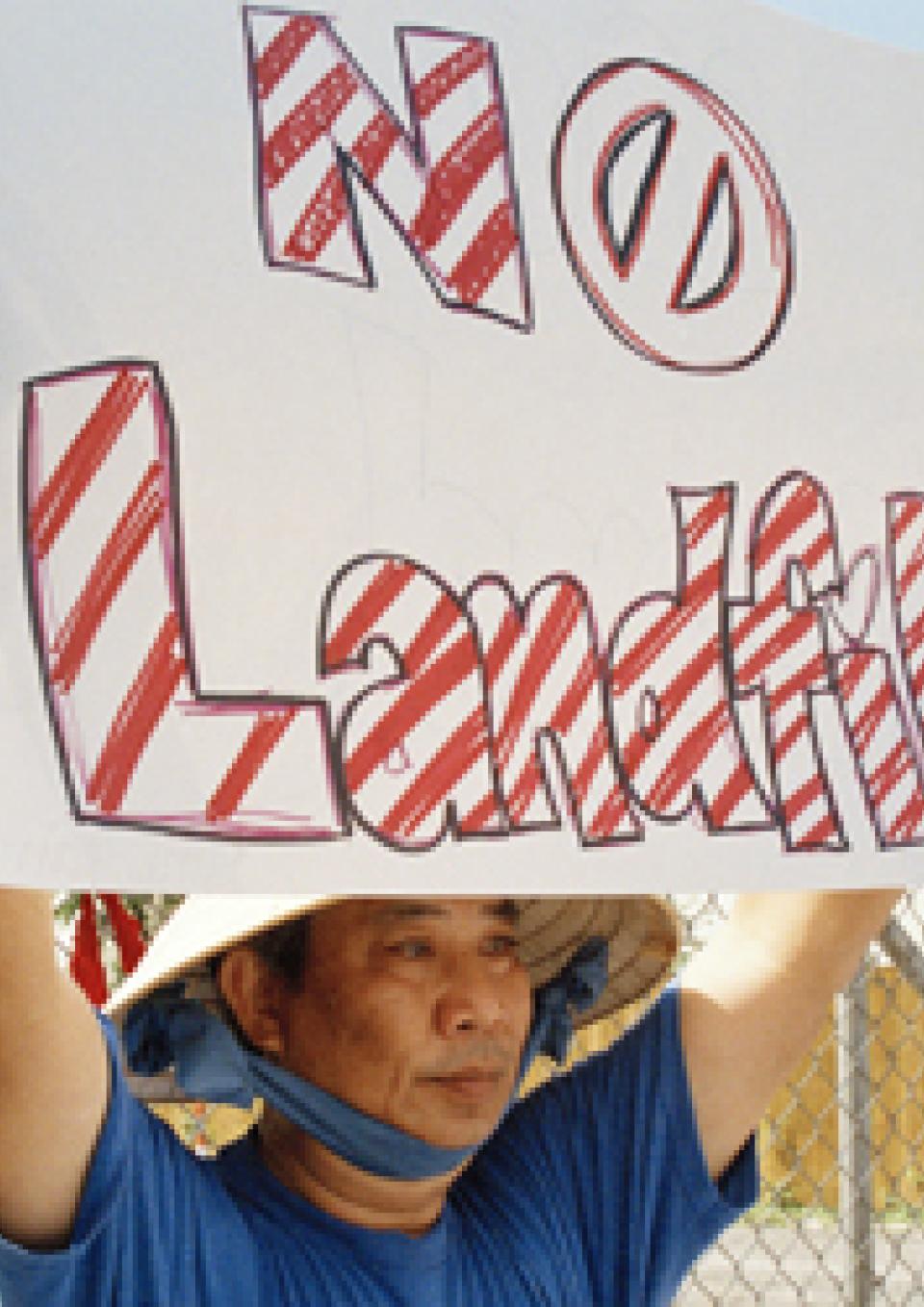  A still from the New Day film A Village Called Versailles. A Vietnamese person holds up a sign that reads “No Landfill” in large red and white striped letters. The “O” in “no” is a circle with a slash through it. They are wearing a conical hat tied under their chin with a large blue ribbon.