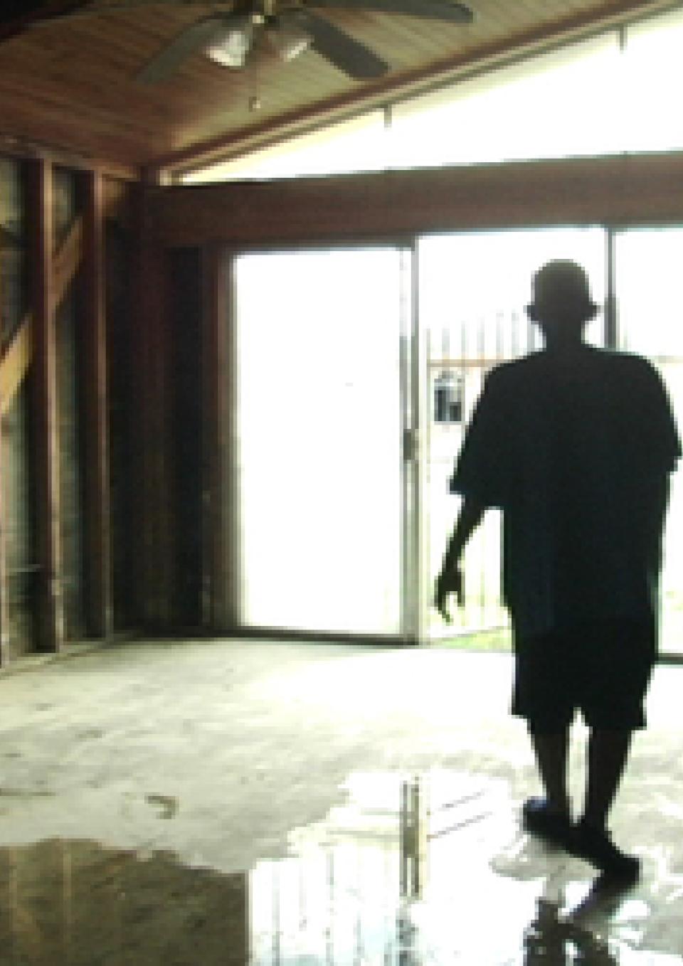 A man stands inside a building where the wall beams are fully exposed and water pools on the cement floor. He is silhouetted against bright windows in the background.