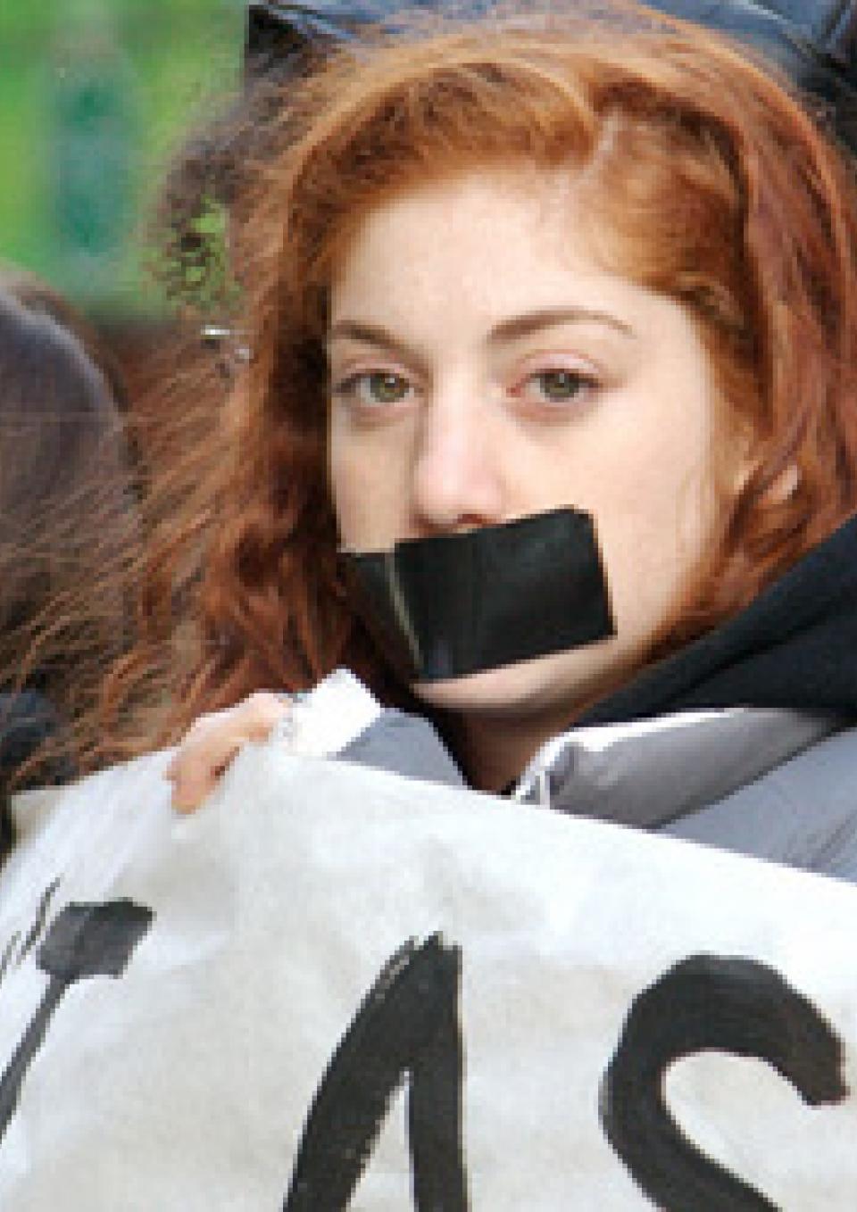 A young girl with red hair stares boldly into the camera in a protest. She has a large black strip of duct tape covering her mouth and she is holding up a protest banner. The other people holding up the banner are blurred in the background.