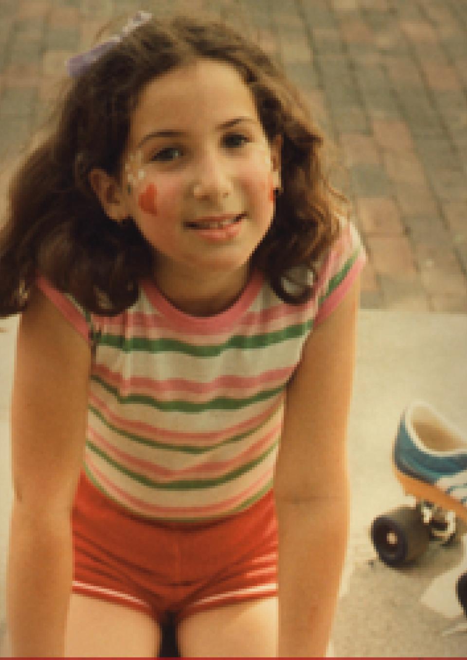 A girl of about ten years old, likely in the early eighties, wearing a shirt with rainbow stripes and red shorts, sitting next to a blue Adidas roller skate with a yellow sole and big black wheels. She has facepaint on her cheeks.