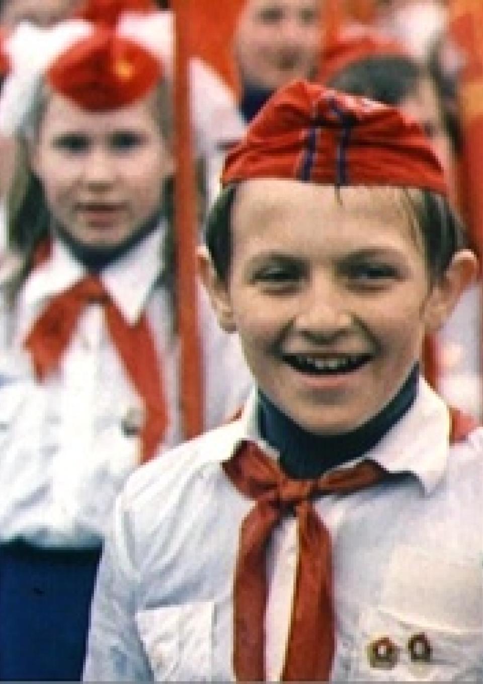 Russian Children stand on guard wearing military outfits which include red hats and scarves, and white shirts with pins on their pockets.