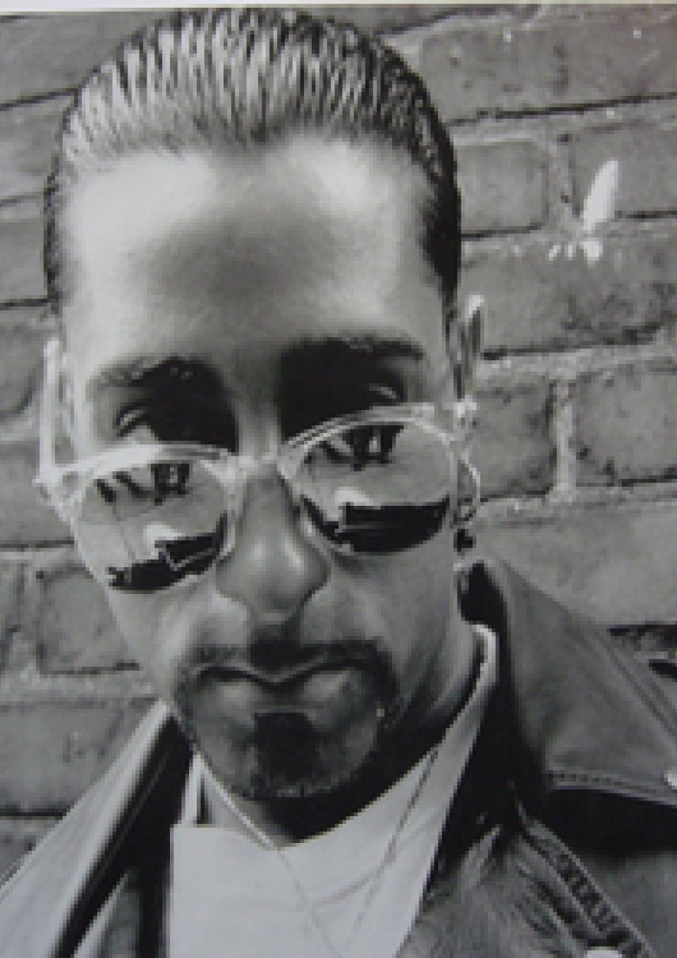 A black and white still from the New Day film Tales from Arab Detroit. A slightly low angle shot of a man with slicked back hair and a leather jacket standing in front of a brick wall. He is looking into the camera, pressing his lips together. He wears sunglasses pushed down on his nose making his eyes partially visible as well as one earring and a necklace.