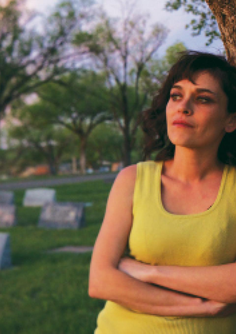 A still from the New Day film Uranium Drive-In. A young woman in a bright yellow sleeveless top crosses her arms and leans against a tree as she looks out thoughtfully into the distance. Behind her are a smattering of graves on bright green grass and more trees.