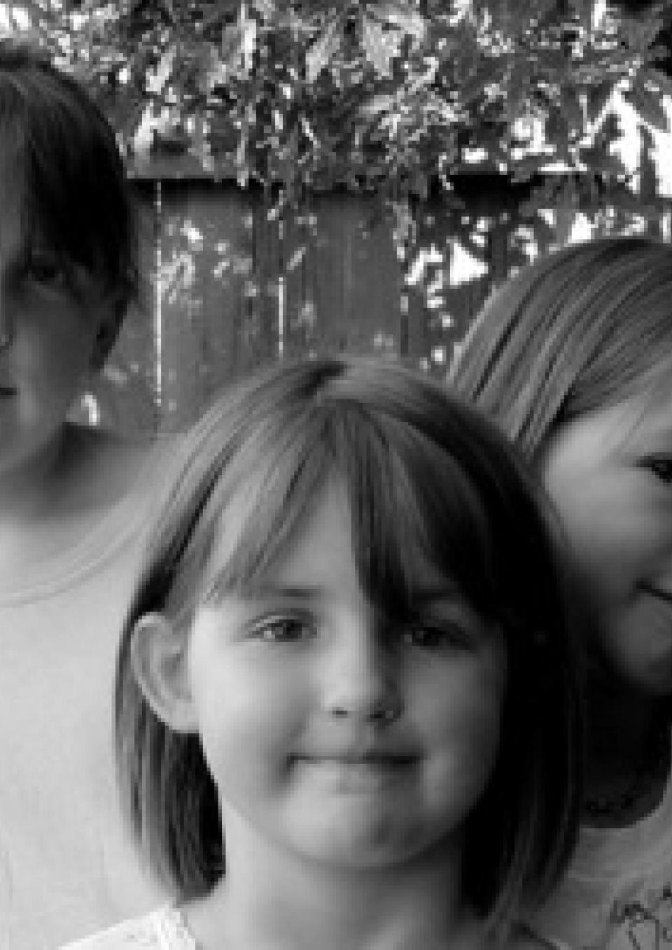 A black and white still from the New Day film No Dumb Questions. Three young girls face toward the camera with close mouthed smiles. The girl on the left peeks out shyly from behind the girl in the center. Behind them is a wooden fence and foliage.