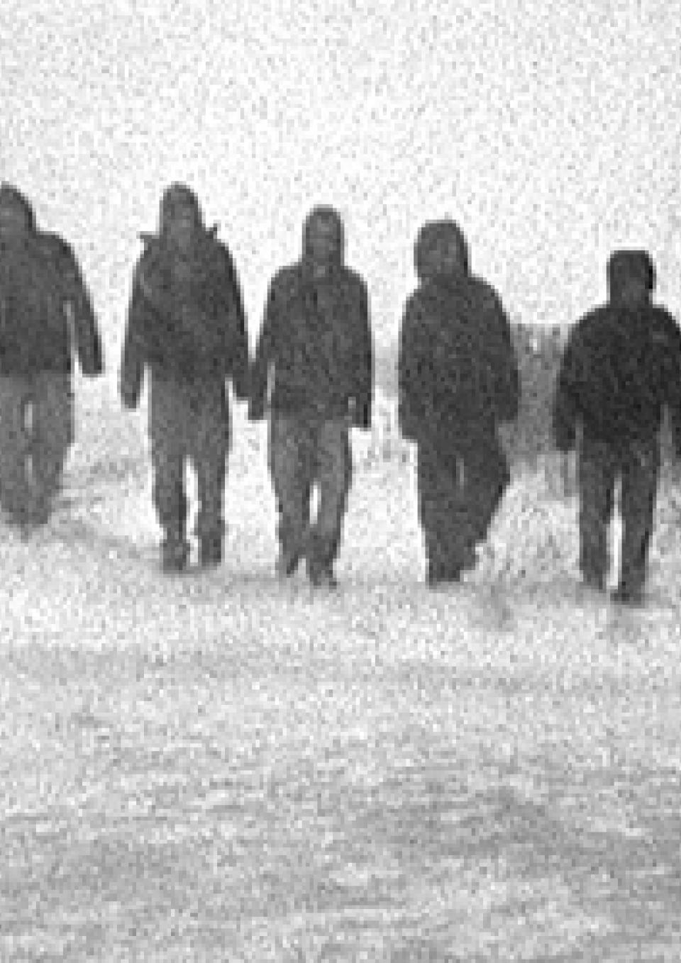 A grainy black and white still from the New Day film Where Soldiers Come From. A row of people shrouded in dark coats and hoods amble down a snowy path. Their surroundings are blurred out with snow but some bushes and trees are visible on the sides of the road.