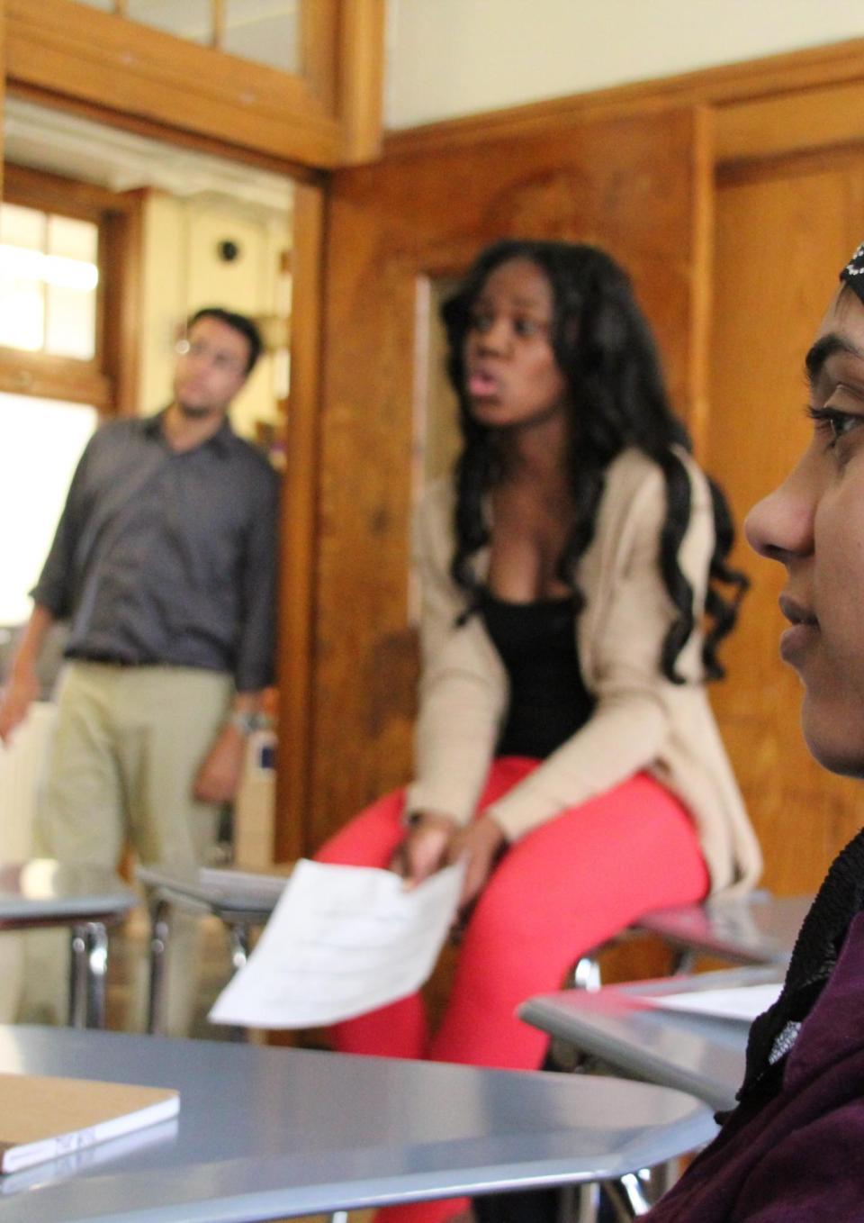 A still from the New Day film I Learn America. A profile of a teenage girl sitting in a classroom wearing a purple shirt and a black head covering. A woman in professional clothing sits on a desk to her right. She holds a piece of paper in her hand and speaks to someone out of the shot. In the background, a man wearing khakis and a button up shirt leans against the wooden door frame. All three are looking at something off camera.