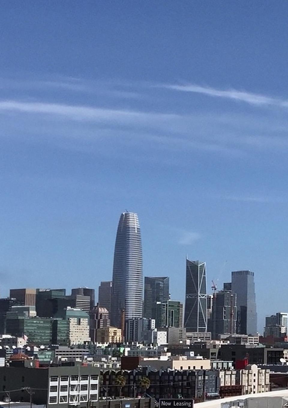 San Francisco skyline with many tall buildings in the background and warehouses in the foreground. In the center is the gigantic Salesforce building, at least a third taller than the tallest of the others, with a curving shape that tapers toward the top. A blue sky with a streak of white clouds takes up most of the frame.
