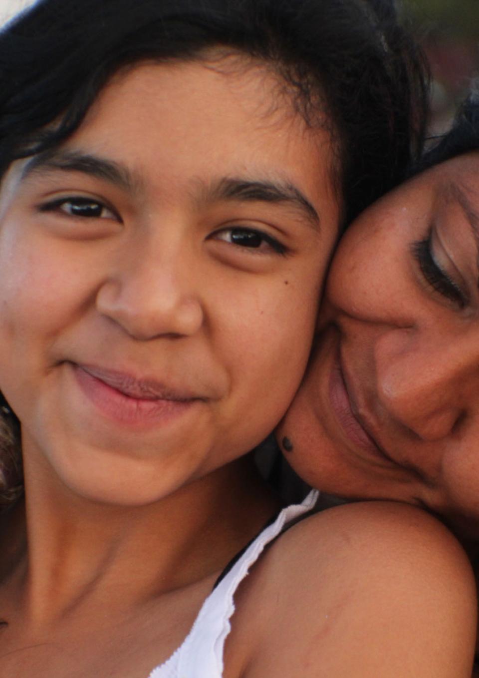 A still from the New Day film Life on the Line. A young Mexican-American girl, Kimberly Torrez smiles sweetly towards the camera. A woman, likely her mother Vanessa Torrez sits behind Kimberly and rests her head on her shoulder. She turns her face into Kimberly’s neck with a loving smile.