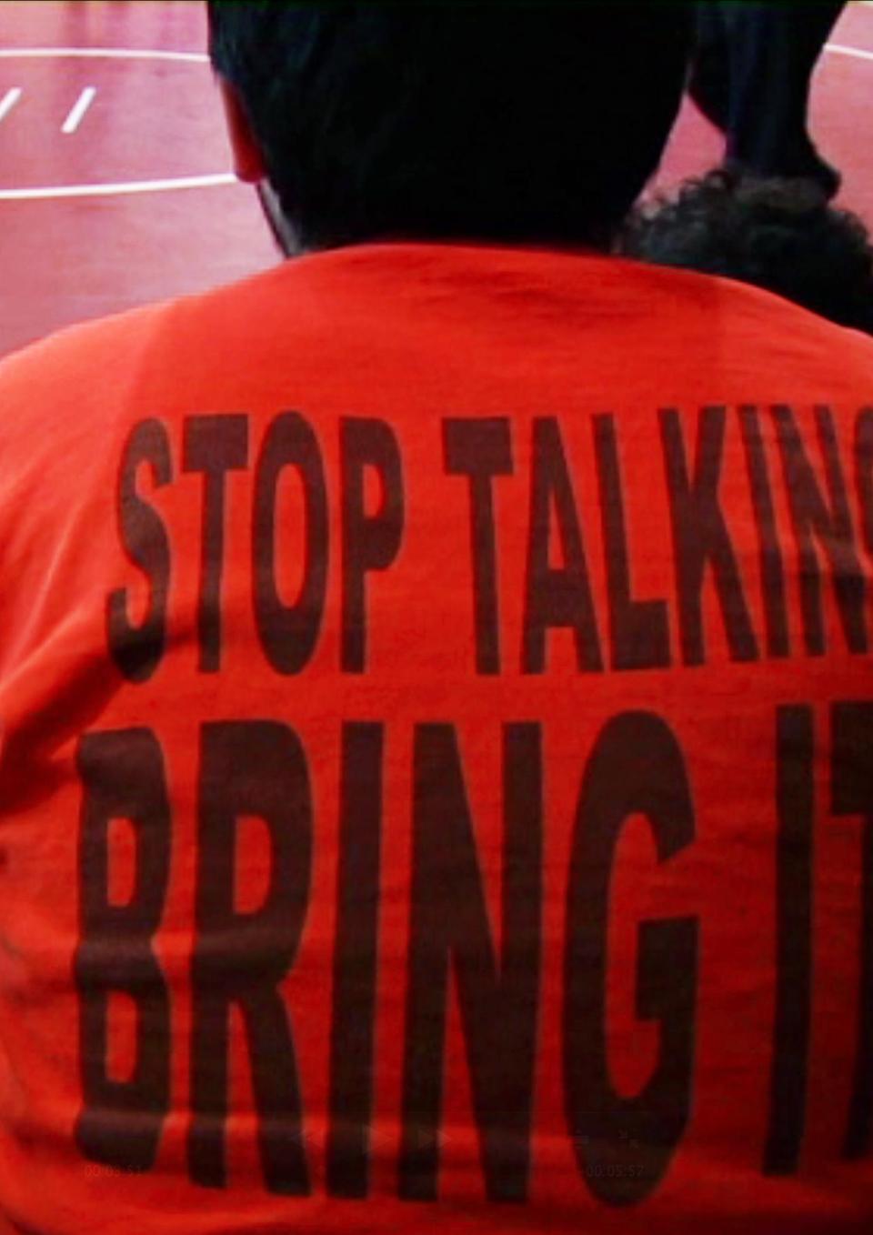 A still from the New Day film Making Noise In Silence. The back of a person sitting on a bleacher. They are wearing a bright orange t-shirt that reads “Stop Talking, Bring It!” in large black letters. The ground is a red gymnasium floor with a wrestling ring.