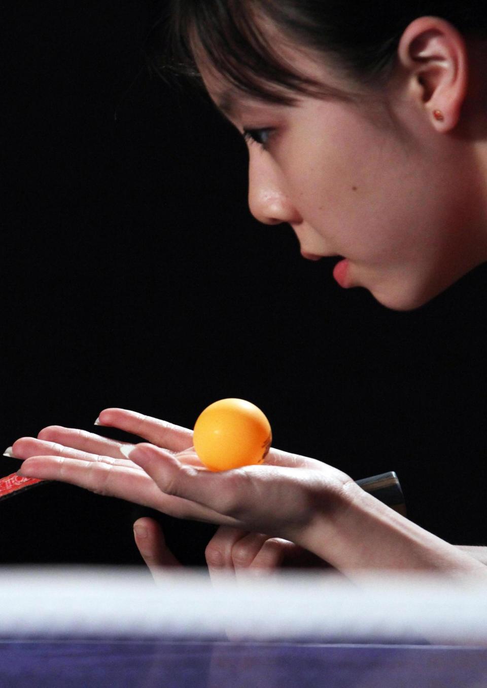 A still from the New Day film Top Spin. An orange table tennis ball rests in Teenage table tennis player Lily Zhang’s open palm. She crouches and looks in the direction of the ball, mouth slightly open in concentration. Her table tennis paddle is visible in her other hand as she prepares to serve the ball.