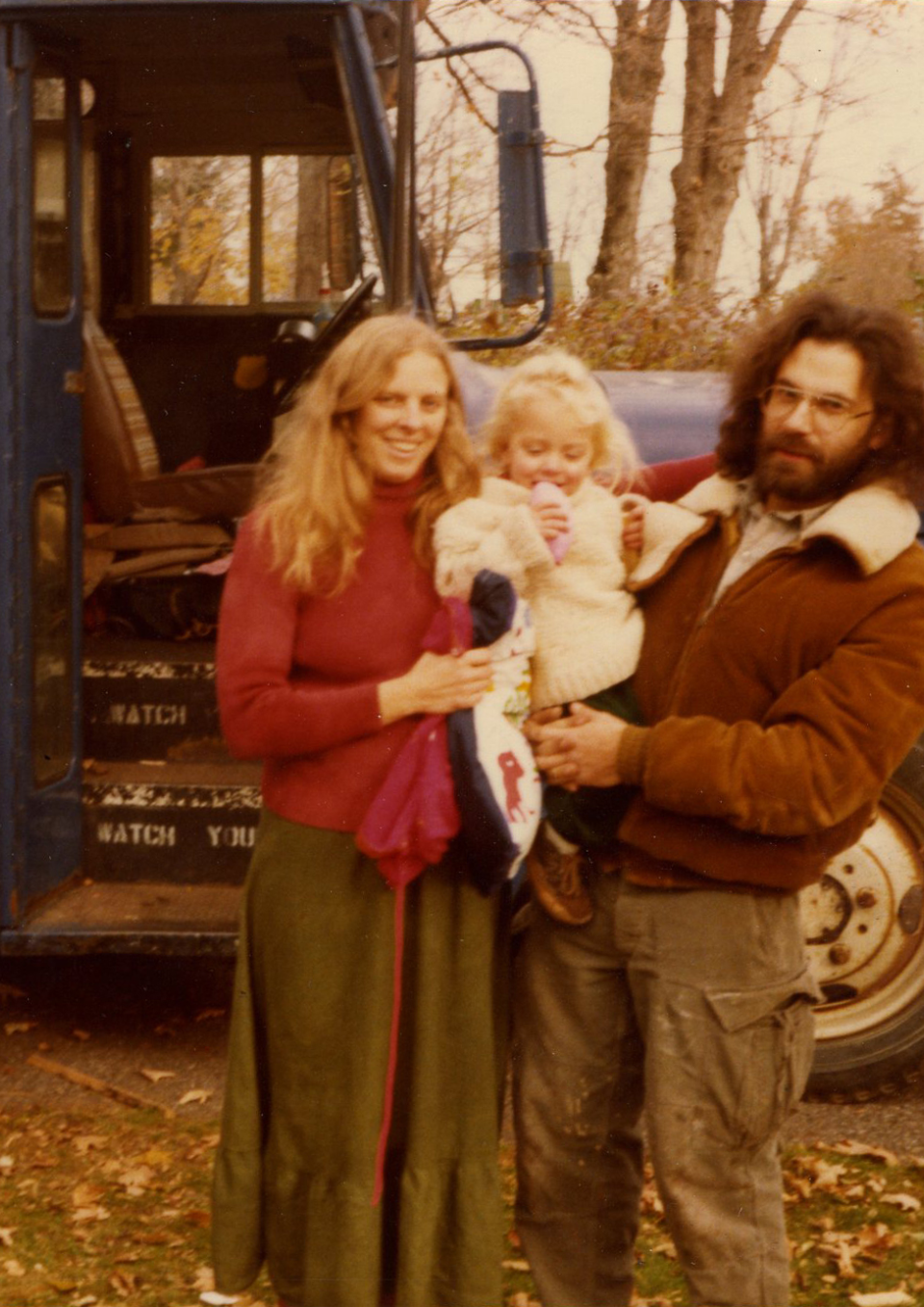 A white woman and man hold a toddler between them. The woman has blond hair and smiles at the camera, the man has long brown hair and a beard and looks unsmiling at the camera. The toddler looks downwards. In the background is a vintage blue school bus.