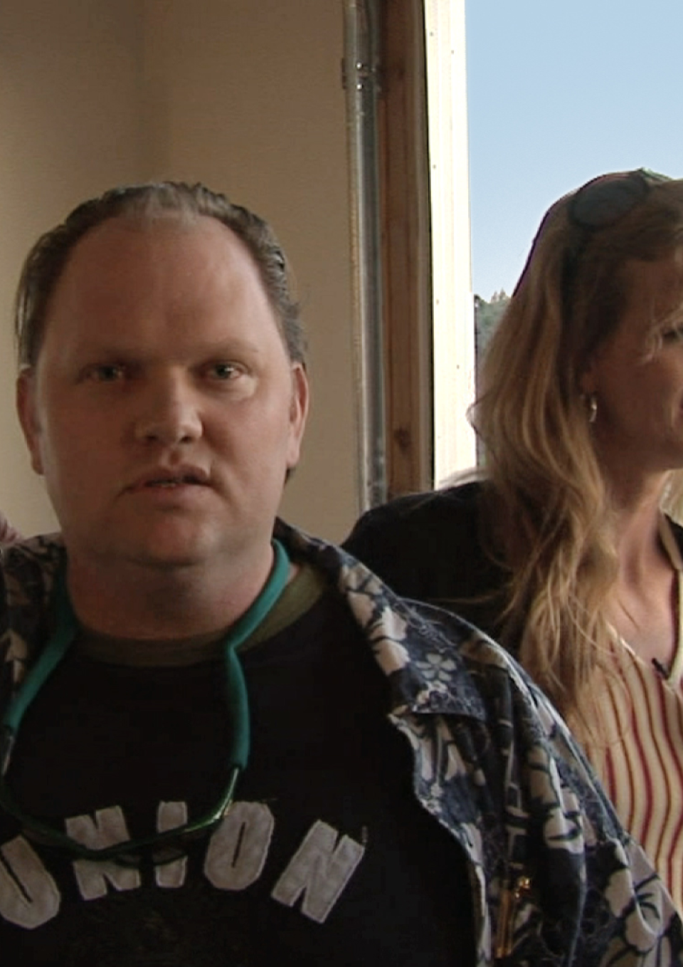 A still from the New Day film Prodigal Sons. A young man faces the camera with a slightly surprised expression as if he wasn’t expecting a picture to be taken. His sister, New Day filmmaker Kimberly Reed rests her hand on his shoulder. She is standing behind him and facing to the side with a drink in her hand. Behind her is a window with trees outside.