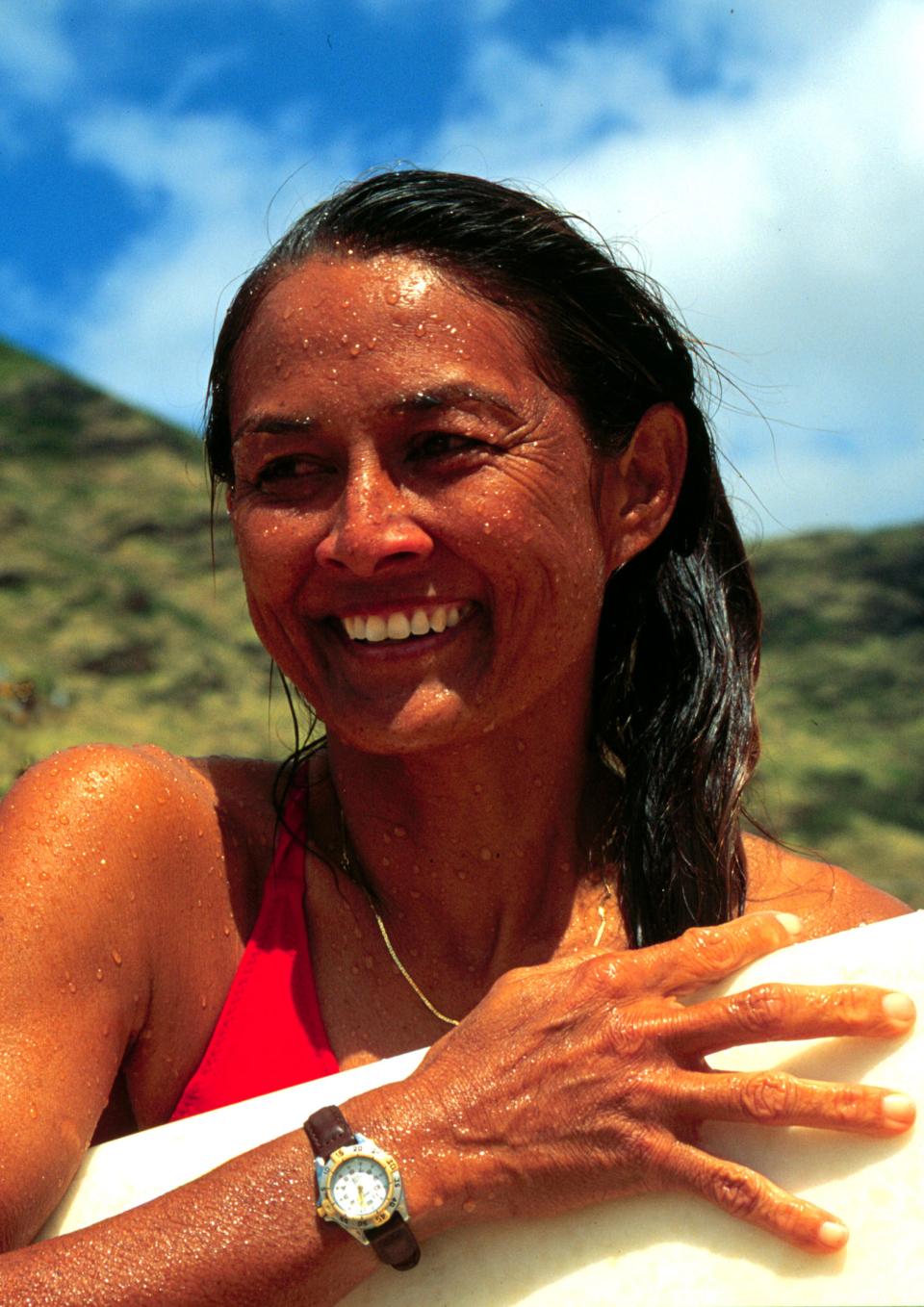 Rell Sunn, a Hawaiian woman, stands outside against a backdrop of lush green mountains and a blue sky with wispy clouds. She is wet with ocean water, her long hair pulled over to the right and draped over her shoulder. She holds a surfboard under her left arm and up against her body and smiles as she looks out to the distance.