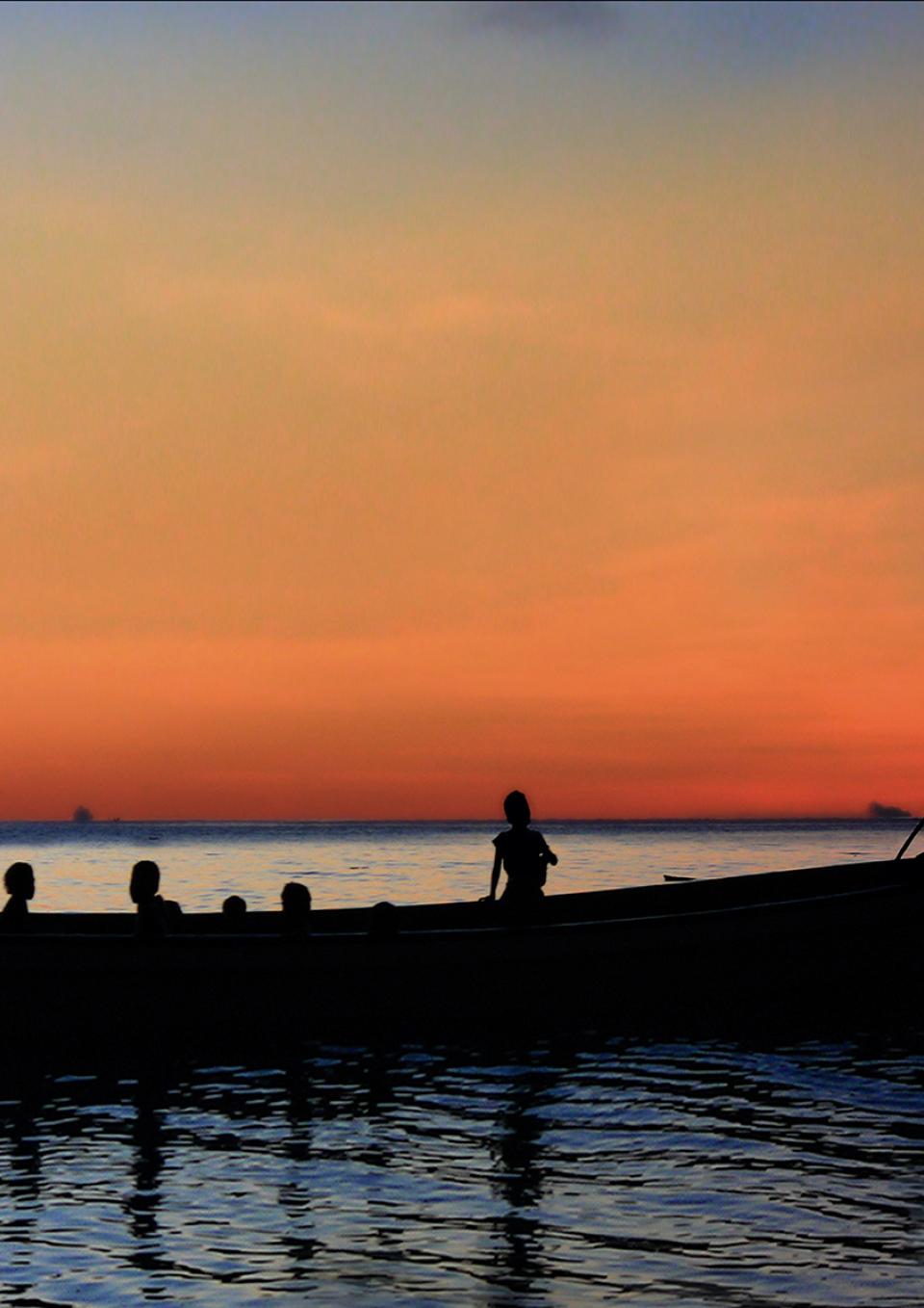 A still from the New Day film Sun Come Up. A silhouette of a large canoe in shiny, calm waters with a deep orange sunset behind. Several people’s heads at different levels show them standing or sitting in the boat. Two people on the right stand at the back of the boat with an oar. A few short buildings disrupt the mostly flat horizon.