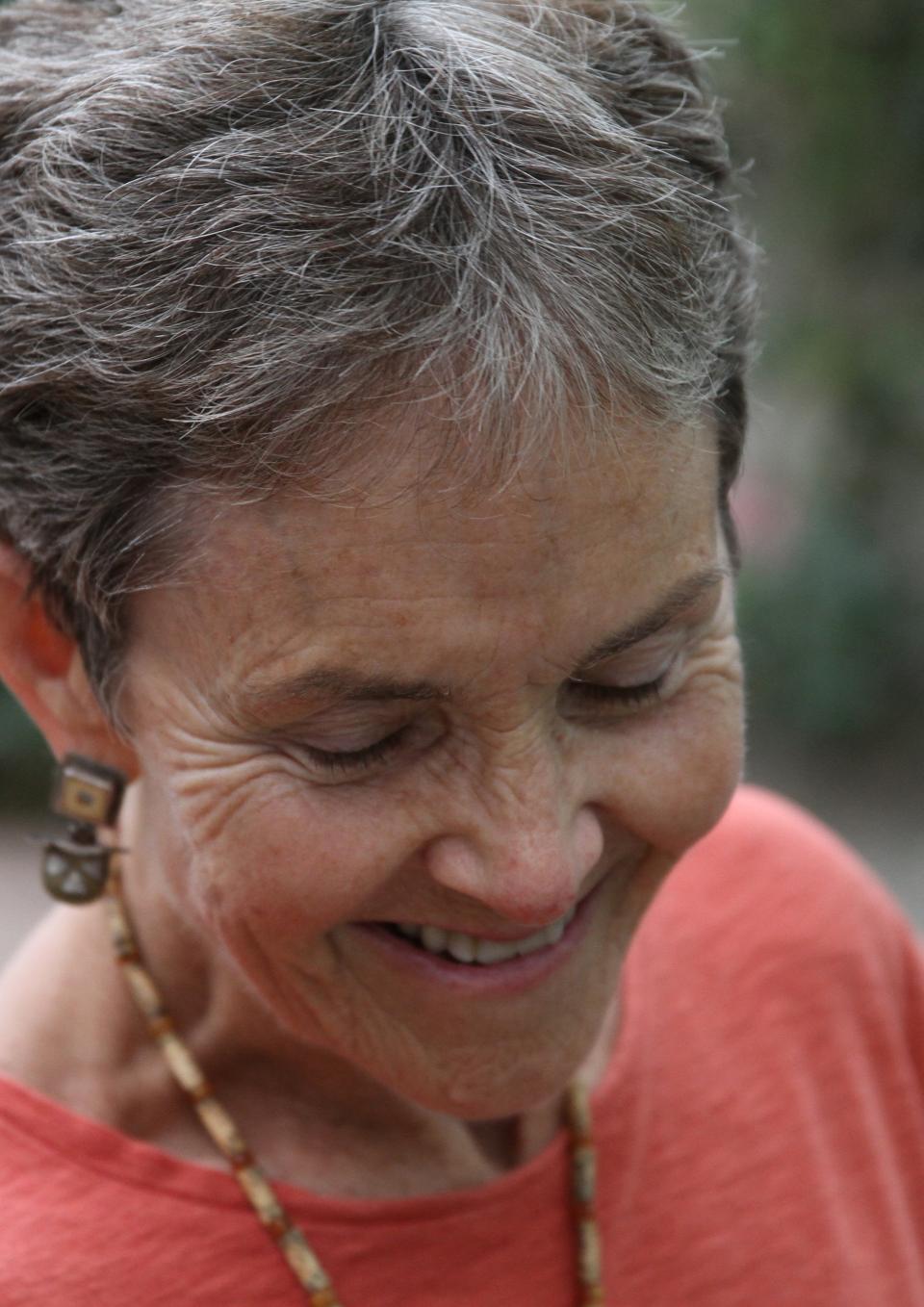 A white woman with short gray hair, smiling, looking down, wearing an orange shirt, a natural colored beaded necklace, and complementary earrings.