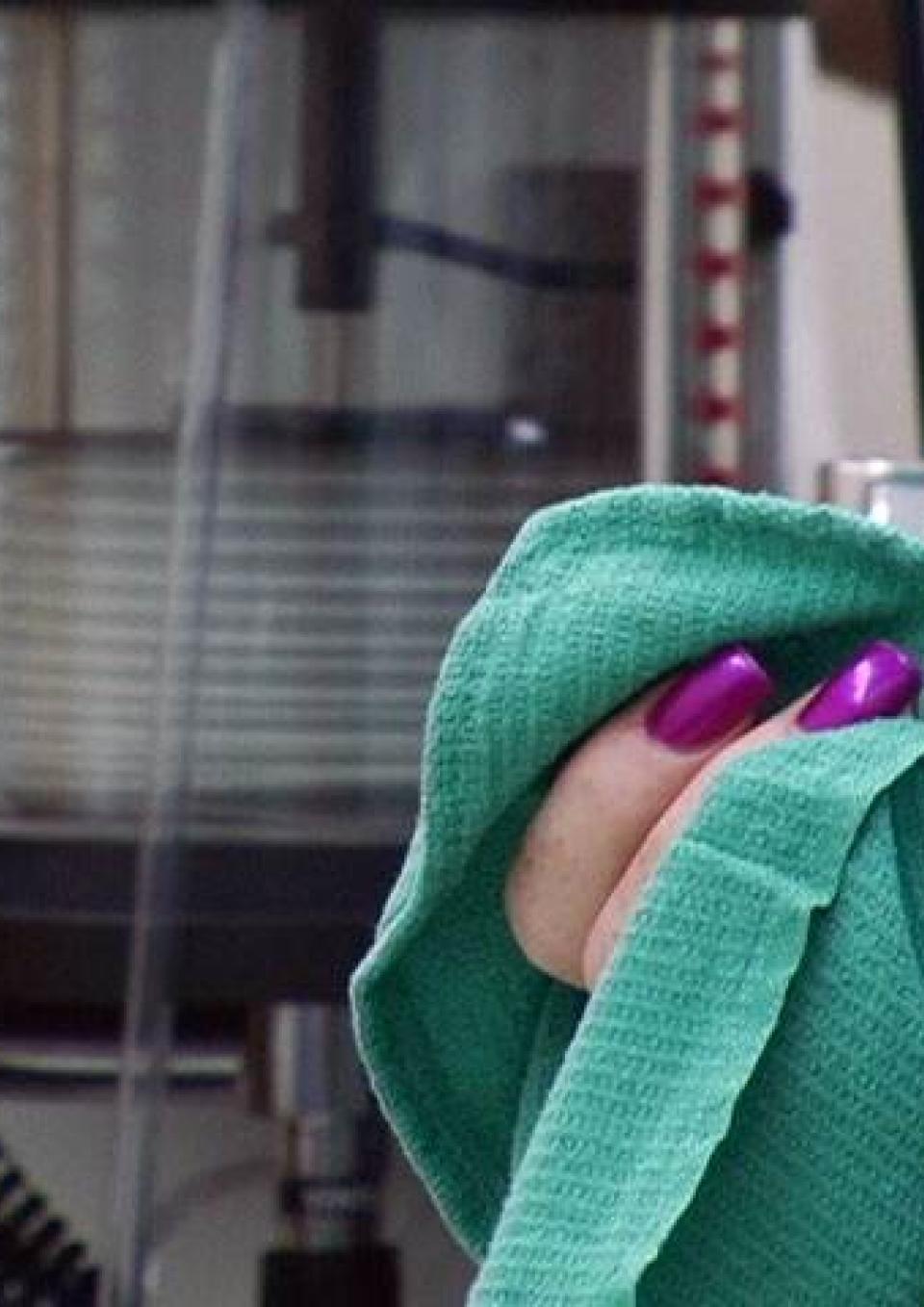 A still from the New Day film Trinidad. A close up of two bright purple nails holding a bright teal cloth. Factory machines fill the blurred background.