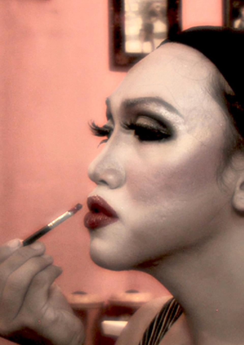 Profile image of a transgender woman in Indonesia. She focuses on her face in the mirror as she paints her lips. She has dramatic eye makeup and her hair pulled back under a cap.