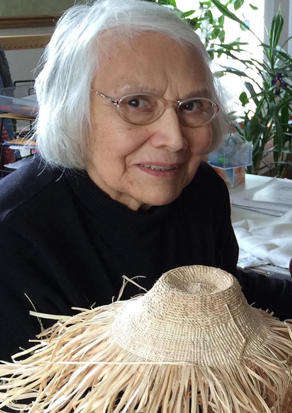 Haida elder and weaver Delores Churchill smiles at the camera. She is wearing glasses and a black top. She holds up a weaving work in progress with the unfinished ends splaying out.