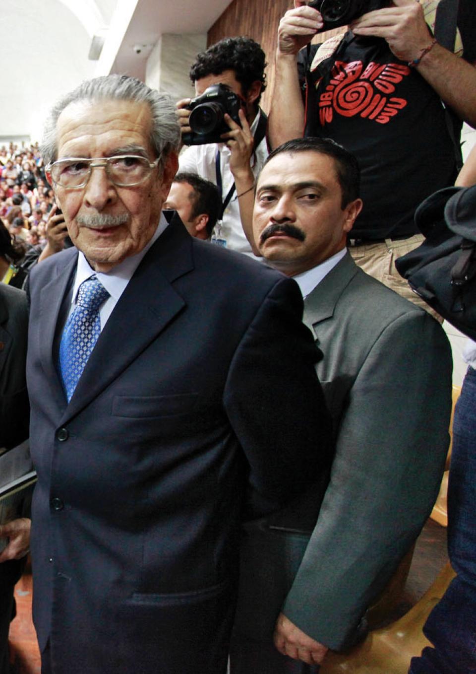 A still from the film “Dictator in the Dock.” An elderly man, General Efrain Rios Montt, and his lawyer are wearing suits and walk, unsmiling, through a crowd of people observing. Several people take pictures of the General as he passes. Another person in a suit stands right behind the General, looking directly into the camera and not smiling.