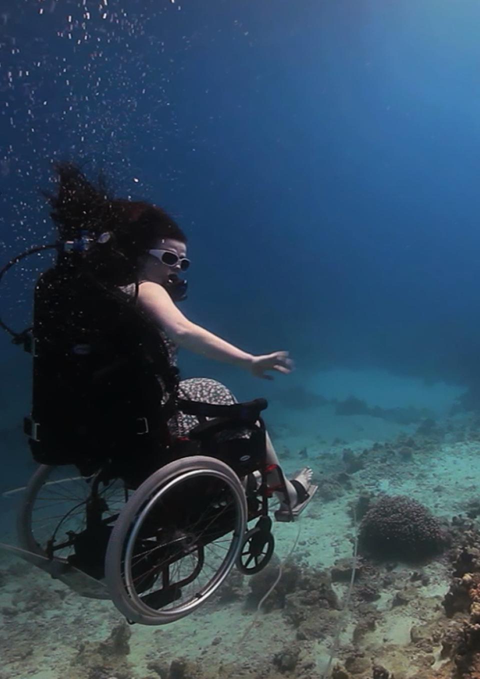 A still from the New Day film Fixed: The Science of Human Enhancement. A woman in a wheelchair is scuba diving. She floats slightly above the ocean floor. Her arms are bare and she does not appear to be wearing full scuba gear other than small goggles. Her hair drifts up in the water and she holds her arms out to the side for balance.