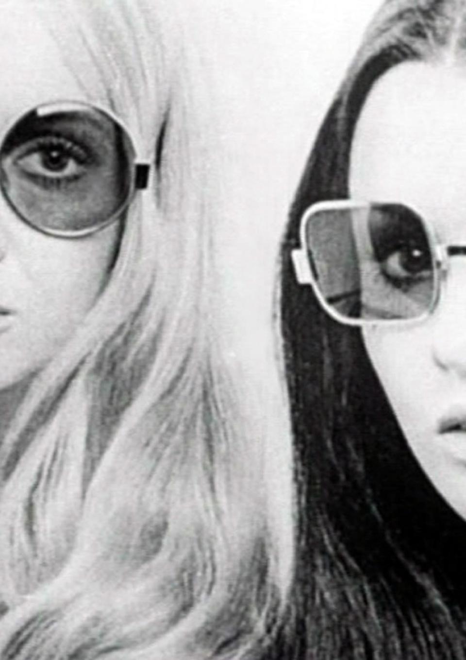 A black and white image from the 1970s of two women, blond and brunette, who stare at the camera, lips parted, with the same still, unsmiling expression. Both wear translucent oversized sunglasses.