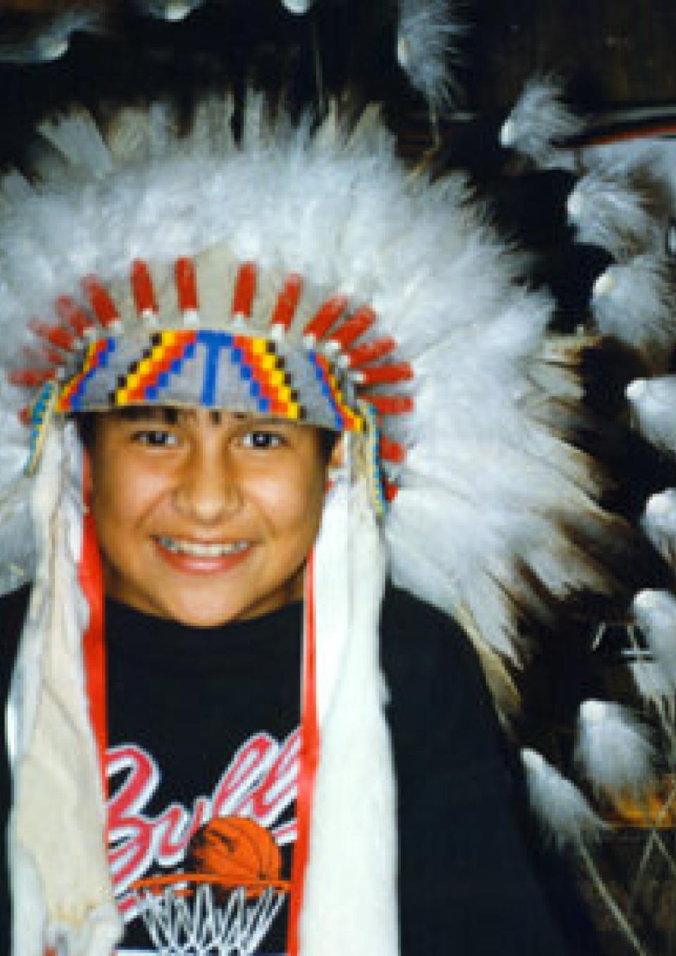 A child with brown skin wears a Native American feather headdress and smiles at the camera. Behind the child on the wall, a blanket and artwork depicting a Native person with long hair.