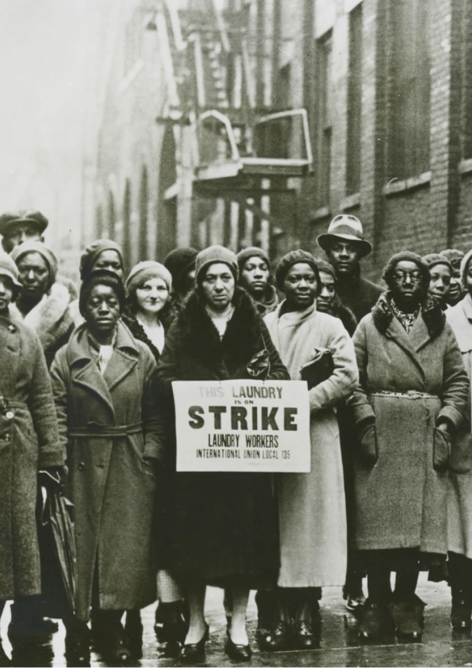 A black and white photo taken in the 1930’s of a group of striking workers who are predominantly Black women. Two women hold signs that say This Laundry is on Strike. Laundry Workers International Union Local 135.