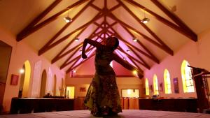 A perfectly centered wide shot of a Black woman dancing in a high-ceilinged sanctuary. She cuts angular lines that evoke the wooden trusses towering above her.