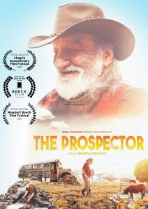 bearded man with black cowboy hat on looking down on school bus in a field with same man walking with a gold pan. The Word "the prospector" are written in gold lettering with 3 film festival laurals on the left side.