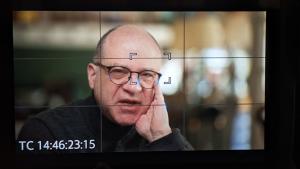 A man wearing glasses and a dark sweater sits before a camera. He is speaking and rests one hand on his face.